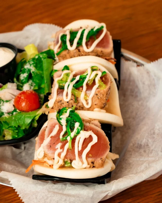Our Sesame Tuna Sliders are the perfect bite! Seared tuna packed into soft steamed bao buns with seaweed salad and a creamy drizzle. Come grab a plate by the water! 🌊😎